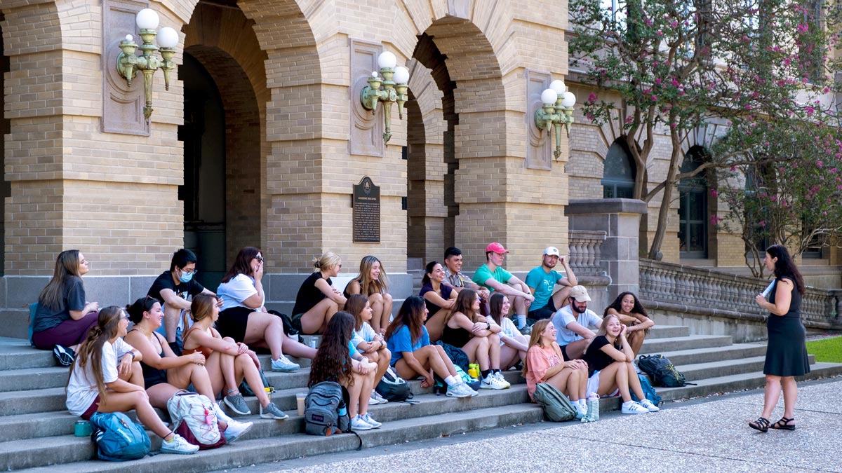 A group of students sits on the steps of a building while an instructor speaks to them